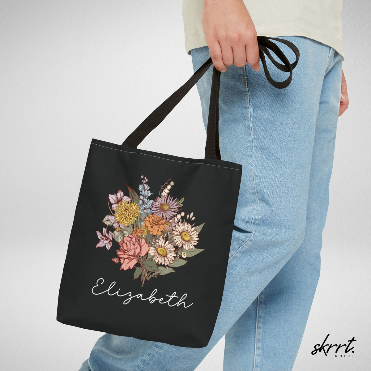 Zwarte tote bag met bloemenboeketprint en naam Elizabeth, hand houdt de hengsels vast.