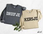 Set: antracietgrijze 'DRUIFJE'-trui en beige 'KERSJE'-trui, plat neergelegd.