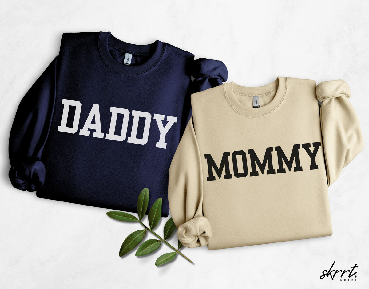 Set van twee sweatshirts: blauw met “DADDY” en beige met “MOMMY”, opgevouwen.